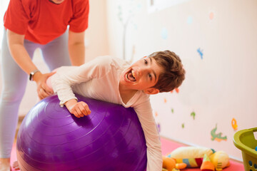 Physical therapist assisting girl with disability lying on fitness ball in rehabilitation centre