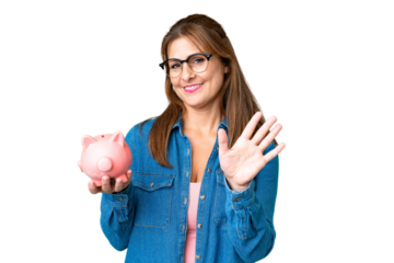 Middle age caucasian woman holding a piggybank over isolated background saluting with hand with happy expression