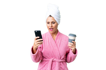 Middle age caucasian woman in a bathrobe over isolated background holding coffee to take away and a mobile