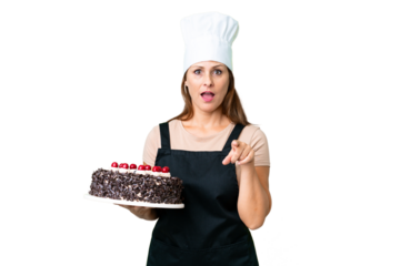Middle age pastry chef woman holding a big cake over isolated background surprised and pointing front