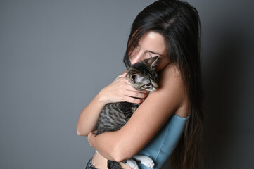 Woman hugging her kitten. Striped kitten lies on on the shoulder of a woman on gray background. 