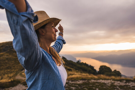 Carefree mature woman with arms raised standing under sky at sunset
