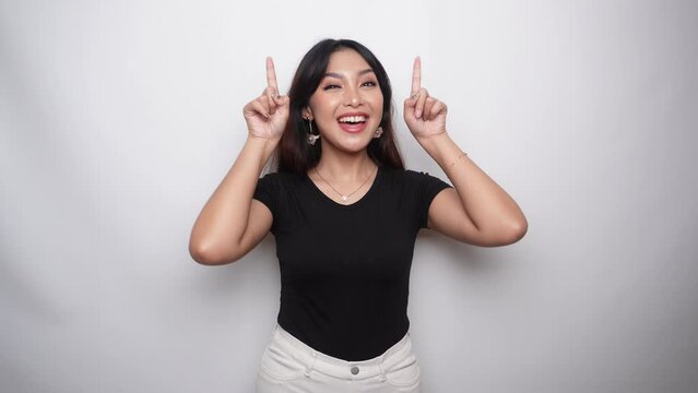 Excited Asian woman dressed in black shirt, pointing at the copy space on top of her, isolated by white background