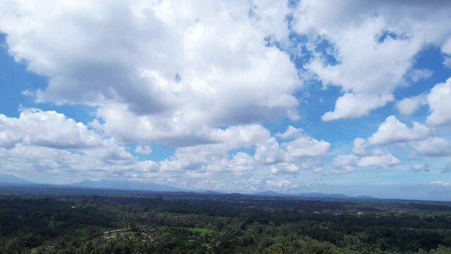 White clouds in the sky, low angle wide shot, aerial camera fly high over Balinese landscape. Smooth slope of very old and huge volcano, land covered with tropical jungles, rice fields and villages