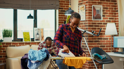 Young modern housewife using heat iron and ironing table, smoothing out creases on clothes. Female person doing household chores for spring cleaning, using laundered clothing.