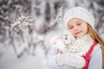 Beautiful little girl with long hair. Little girl in the winter forest. Girl in a hat. Girl in a Zara sheepskin coat. A little girl hugs a toy hare. Winter. Snow. Tender baby. Baby portrait.