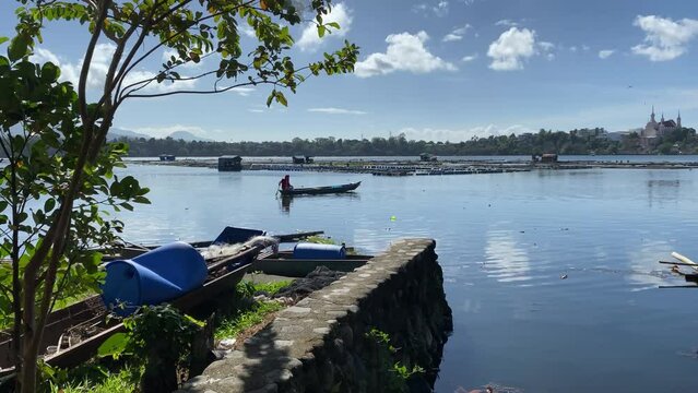 Broke down wooden fishing boat abandoned on the stone riprapped polluted lake breakwater, boatman rowing boat in the background