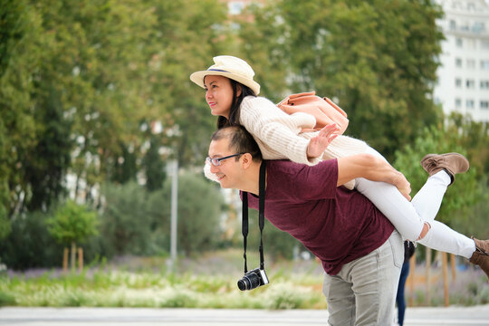 Multiracial Couple Of Tourists Piggyback With Spread Arms Like Plane Wings. Sightseeing In Madrid, Spain.