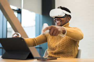 An excited black man exploring virtual reality using VR glasses and controllers in an office,...