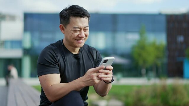 Smiling adult asian man using a smartphone while sitting on a bench against the background of a modern city building. A handsome male is reading a pleasant message, texting with a friend or girlfriend