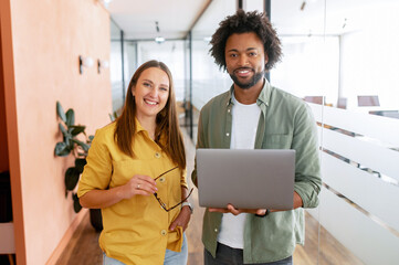 Two happy diverse colleagues African American man and Caucasian woman looking at camera standing in office lobby hall, the man is holding laptop - obrazy, fototapety, plakaty