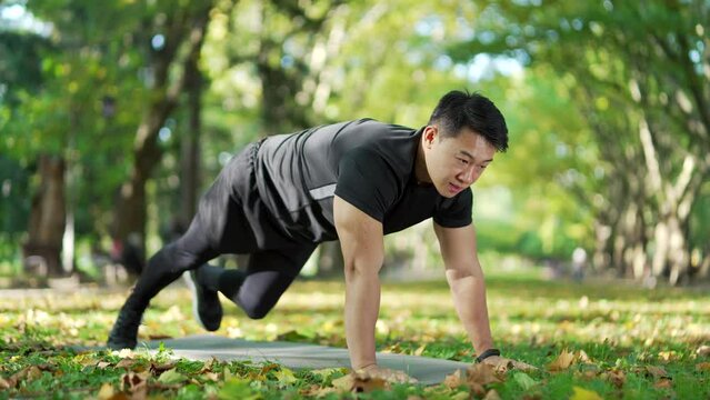 Athletic asian man doing gymnastics on a mat in an urban city park. A handsome male in a sports suit is busy exercising outside. A healthy sportsman trains his abdominal muscles in a push-up position