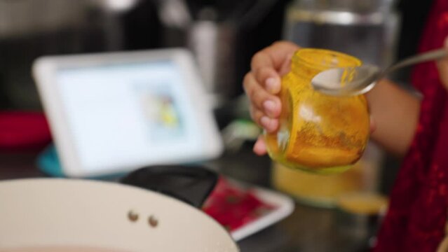 South Asian woman's hands putting a spoonful of turmeric in a pot of curry.