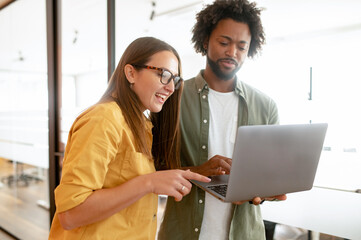 Two young happy colleague using laptop in office hallway and communicating while working together in coworking space, sharing new ideas, discussing project strategy