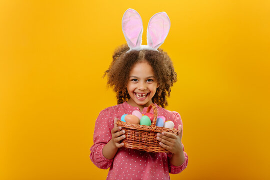 A girl is holding a basket full of Easter colored eggs on a yellow background