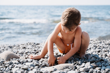 Fototapeta na wymiar A cute boy is relaxing on the beach, playing with sea pebbles. Outdoor activities on the seashore