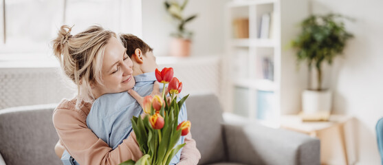 Cute boy sitting on the sofa with mom and giving a bouquet of tulips to her. - obrazy, fototapety, plakaty