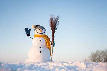 Funny snowman in knitted hat and yellow scalf with broom on snowy field. Blue sky on background