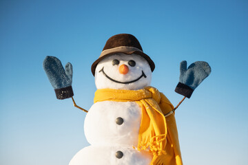 Funny snowman in brown hat and yellow scalf with hands up on blue sky background