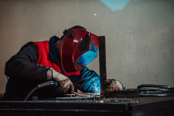 Profesional welder in protective uniform and mask welding metal pipe on the industrial table with...