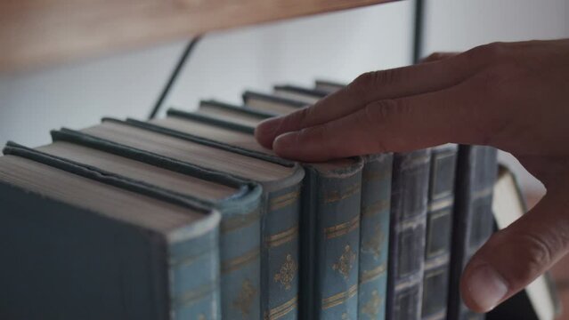 A man takes a book from a row of books on a bookshelf. Old books in leather binding. Close up on the hand and books.