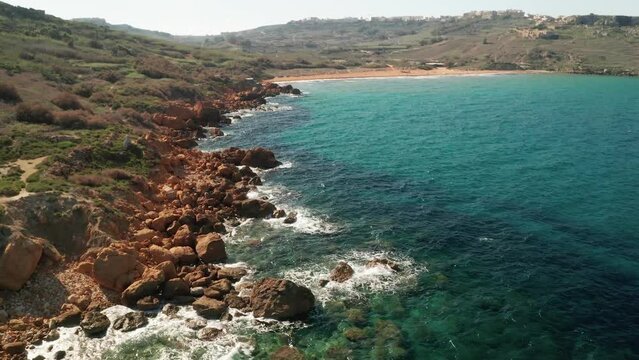 Aerial tilt down reveal shot of the shore near Rampla Beach with beautiful red rocks and clear turquoise water and waves crushing on rocks. Island of Gozo, Malta. Camera is tilting down slowly.