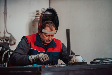 Profesional welder in protective uniform and mask welding metal pipe on the industrial table with...