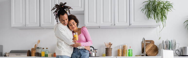 Smiling african american couple with orange juice hugging in kitchen, banner.