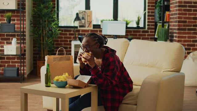 Happy woman preparing cheeseburger and fries, eating fast food takeout meal from delivery paperbag on sofa. Modern girl enjoying dinner wth burger and beer, watching movie on television.
