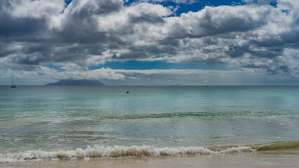 The waves of the turquoise ocean roll ashore and foam on the sandy beach. In the distance - the silhouette of a yacht. The island on the horizon is hiding in the clouds. Blue sky. Seychelles. Mahe.