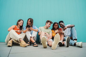 Fototapeta Multiracial group of young friends enjoying and smiling using their mobile phone app sitting at teal wall. Diverse teenagers having fun sharing messages with each other on cellphone at blue clor obraz