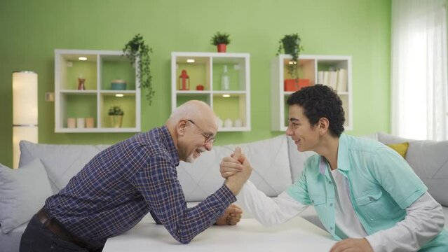Funny grandfather and strong grandchild are arm wrestling.
Old grandfather and grandson arm wrestle at home.
