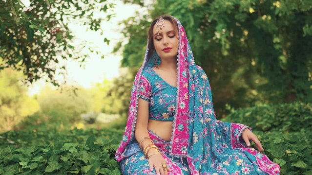 fantasy woman sits on green grass ivy, enjoy summer green nature, tropical garden park. National India blue pink dress saree, scarf on head. Silver teak tiara. Indin girl fashion model beauty face. 4k