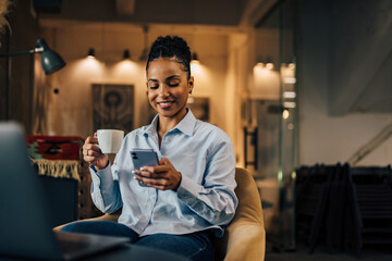 Happy African businesswoman using a phone and drinking a coffee.