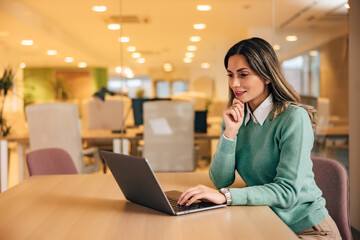 Smiling businesswoman typing on a laptop, sitting at the office, elegantly dressed.