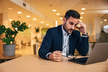 Worried man boss looking at the laptop, holding glasses, sitting at the workplace.