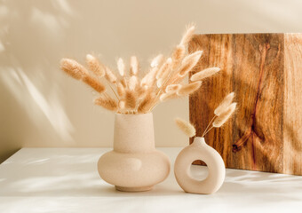 Set of ceramic vase with lagurus grass and wooden stand at the background, warm sunlight shadows, cozy home decoration. Scandinavian interior
