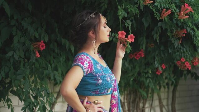 fantasy awoman model posing in image Indian princess. Adult girl holds in hand enjoy beauty aroma, smell blooming red orange flower Kampsis bush green branch tree summer nature. Pink blue saree dress 
