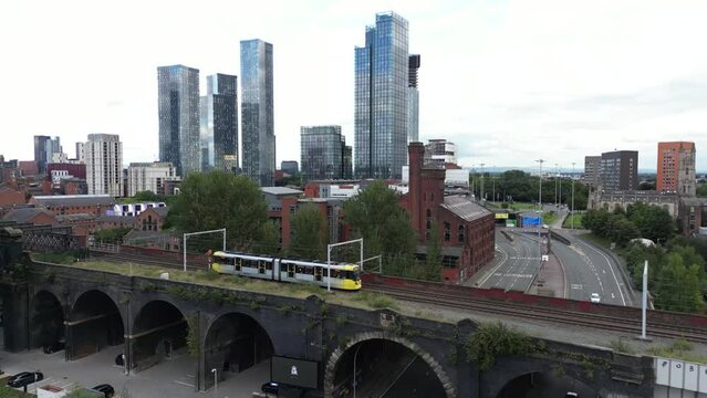 Aerial drone flight alongside a tram on a bridge showing a background of Manchester City Centre skyscrapers and apartment buildings