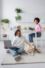 Smiling african american couple with laptop looking at labrador on carpet at home.