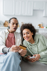 Smiling african american woman using cellphone while boyfriend holding credit card on couch.