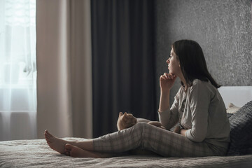 A sad young woman in pajamas with seasonal affective disorder sits alone on the bed and looks out...