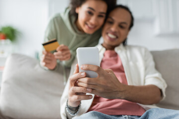 Blurred african american couple using smartphone and credit card during online shopping at home.