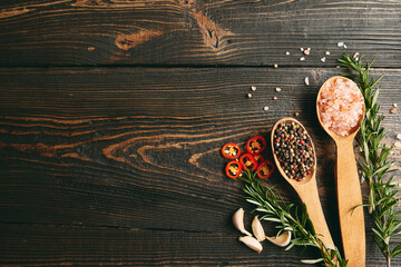 Top view of mixed peppercorns and sea salt in wooden spoons, fresh rosemary and garlic on dark wooden background