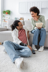 Positive african american couple with popcorn and remote controller talking at home.