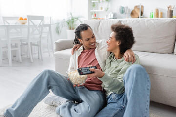 Positive african american couple with popcorn hugging while watching movie at home.