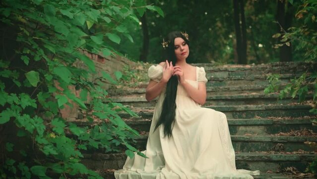 Fantasy medieval woman in white vintage dress. Brunette girl, black long hair, gold wreath. Lady sitting dreaming on staircase, eyes closed, enjoying nature, listening sounds of music. Old retro style