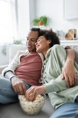 Smiling african american couple hugging near blurred popcorn on couch.