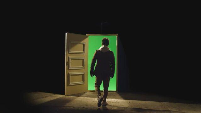 Back view of a man walking in front of an open door with darkness behind . Footage of businessman walking on floor near door with green screen . Concept of anonymous,  business dreams or teleportation