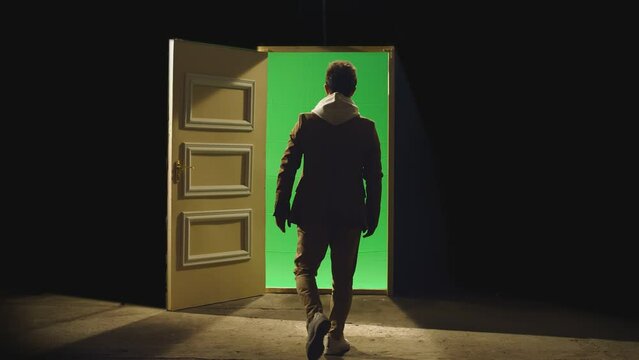 Back view of a man walking in front of an open door with darkness behind . Footage of businessman walking on floor near door with green screen . Concept of anonymous,  business dreams or teleportation